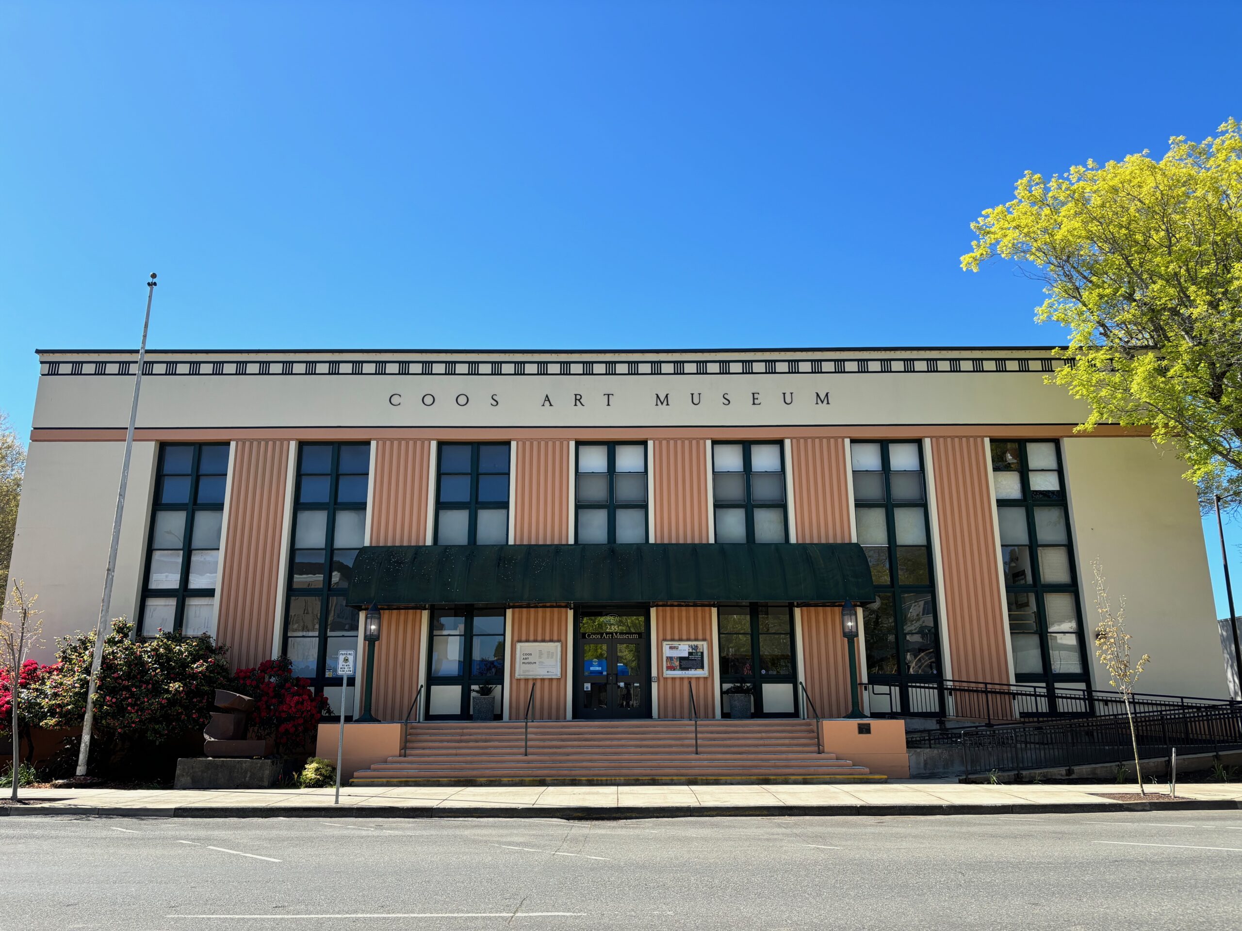 The image shows the front of Coos Art Museum, a rectangular building with a cream-colored façade and vertical peach columns flanking tall, dark-framed windows. The museum's name, "COOS ART MUSEUM," is displayed prominently at the top. A green awning covers the entrance, and a few blooming bushes with red flowers sit on the left side of the building. The sky is clear and blue, and a large tree on the right adds greenery to the scene.